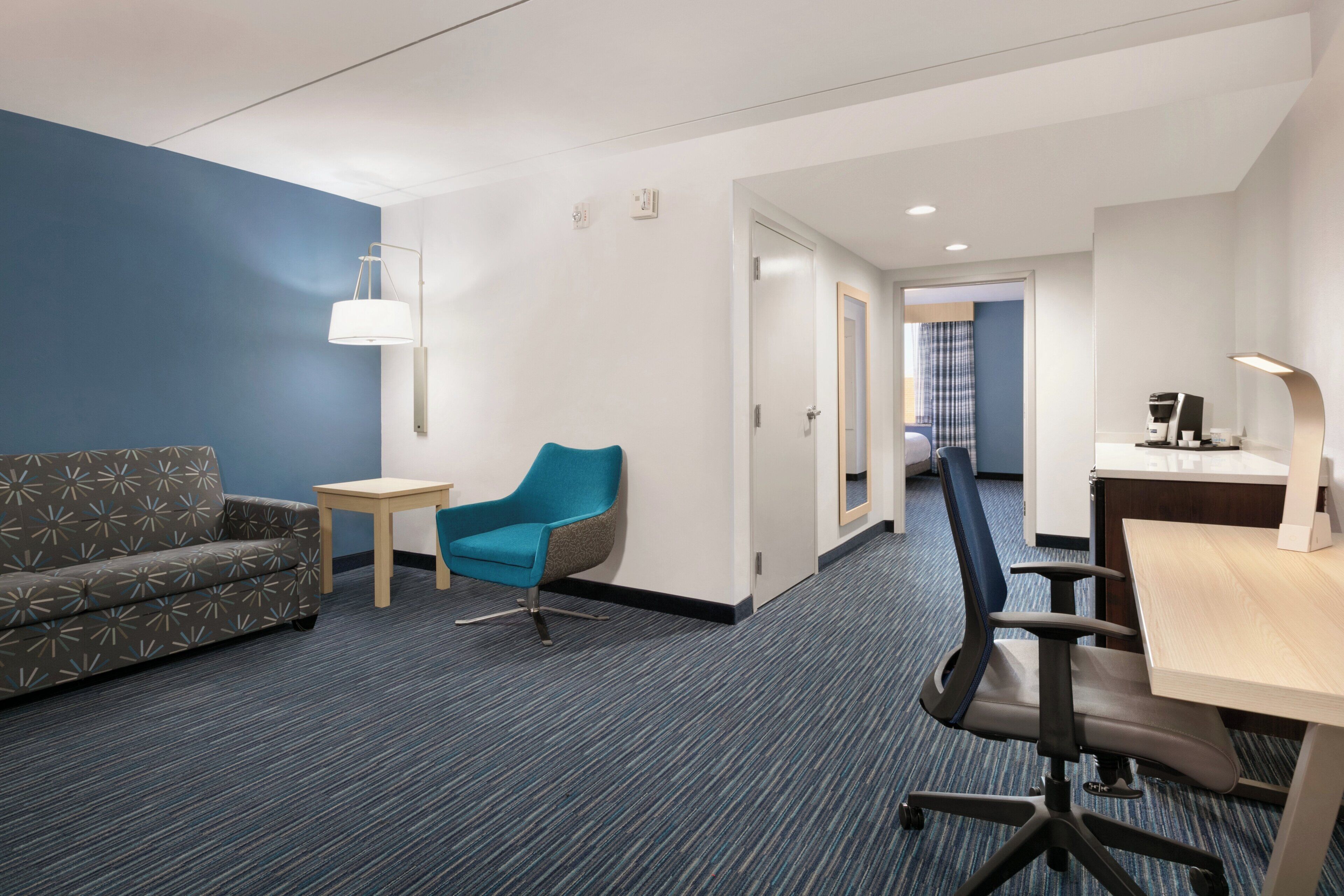 A modern hotel room with a blue accent wall, a patterned sofa, a blue armchair, a small table, a desk with a computer, and a coffee machine.