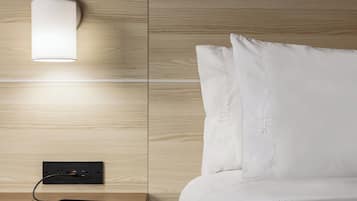 A neatly made bed with white bedding, a bedside table with a lamp, and a power outlet with a phone on it.