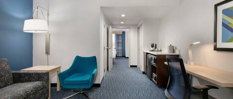 A hotel room with a sofa, a blue armchair, a desk, a chair, and a lamp.