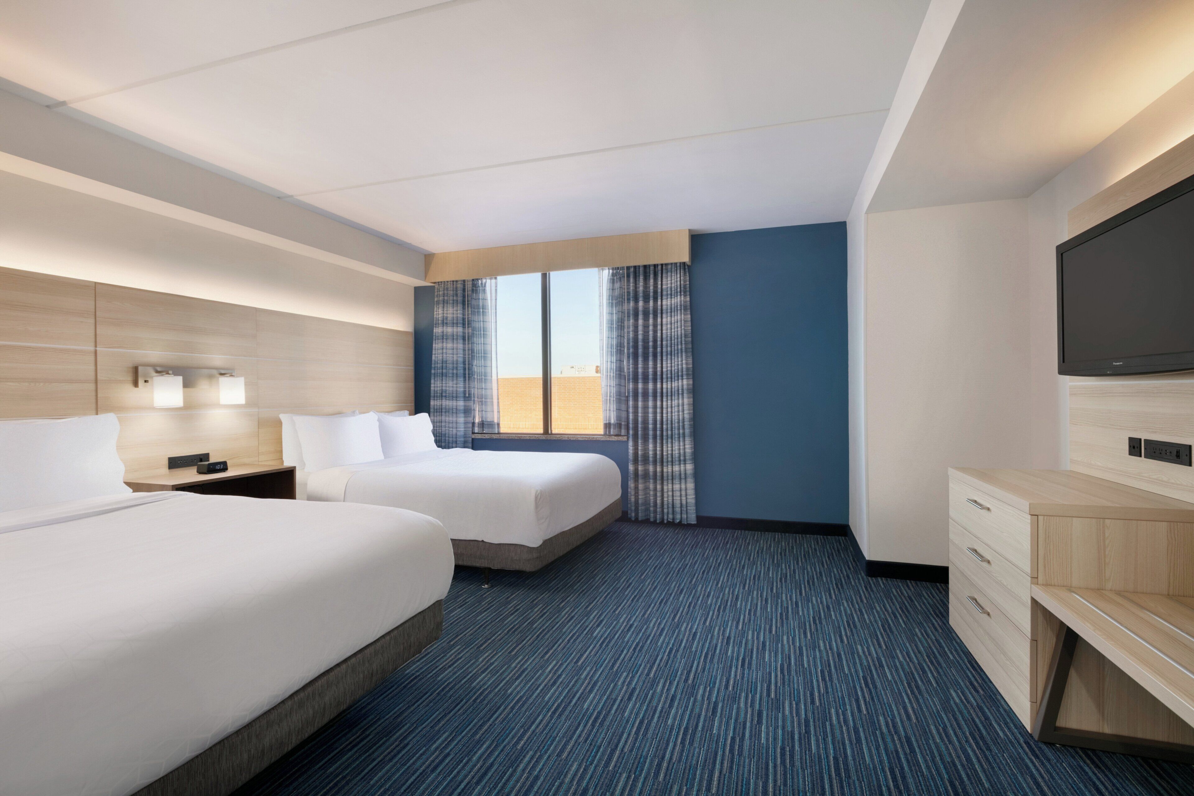 A hotel room with two beds, a flat-screen TV, a desk, and a large window with curtains.