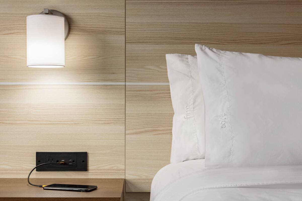 A neatly made bed with white bedding, a bedside table with a lamp, and a power outlet with a charging cable.
