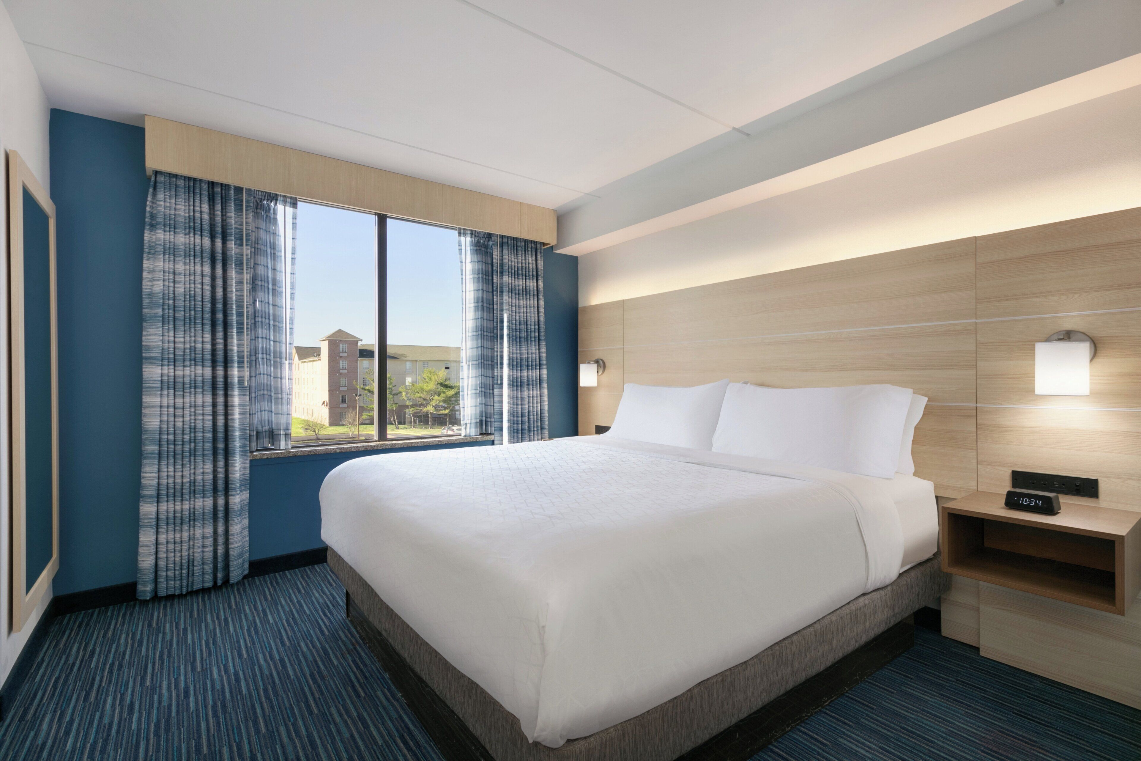 A hotel room with a large bed, a nightstand, a clock, and a window with curtains.