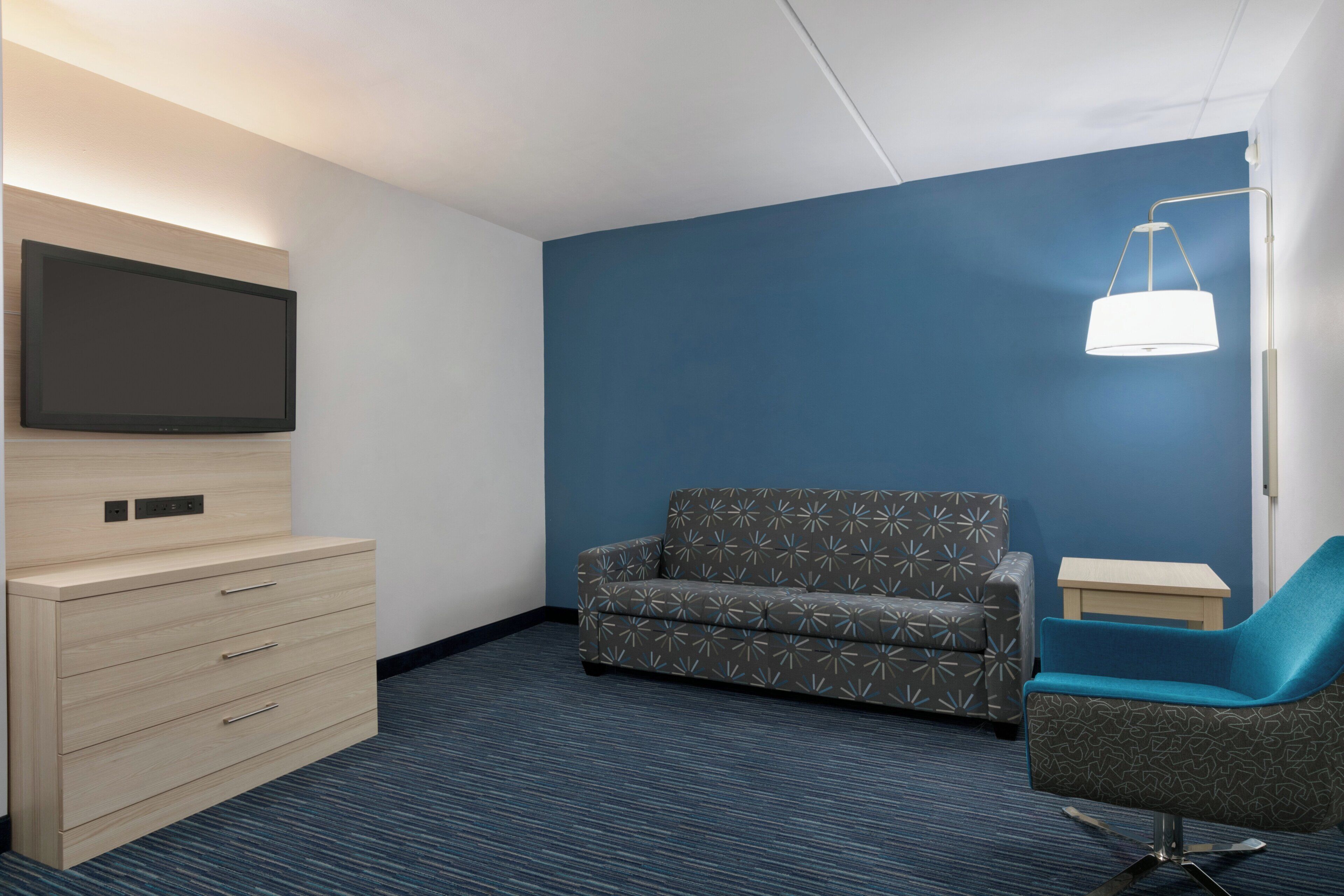 Habitación de hotel con televisión de pantalla plana montada en un mueble de madera, un sofá, una silla y una mesita de noche.