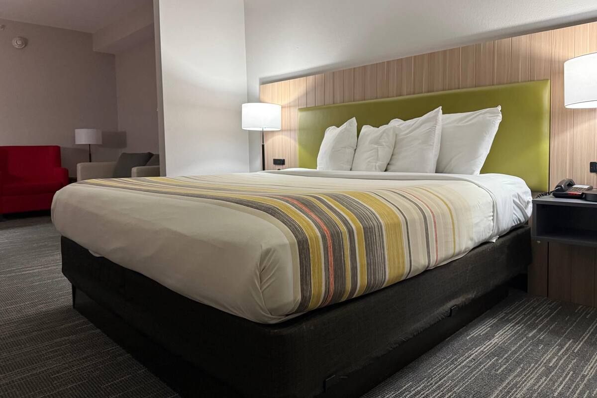 A hotel room with a large bed, a red chair, a desk with a lamp, and a phone.