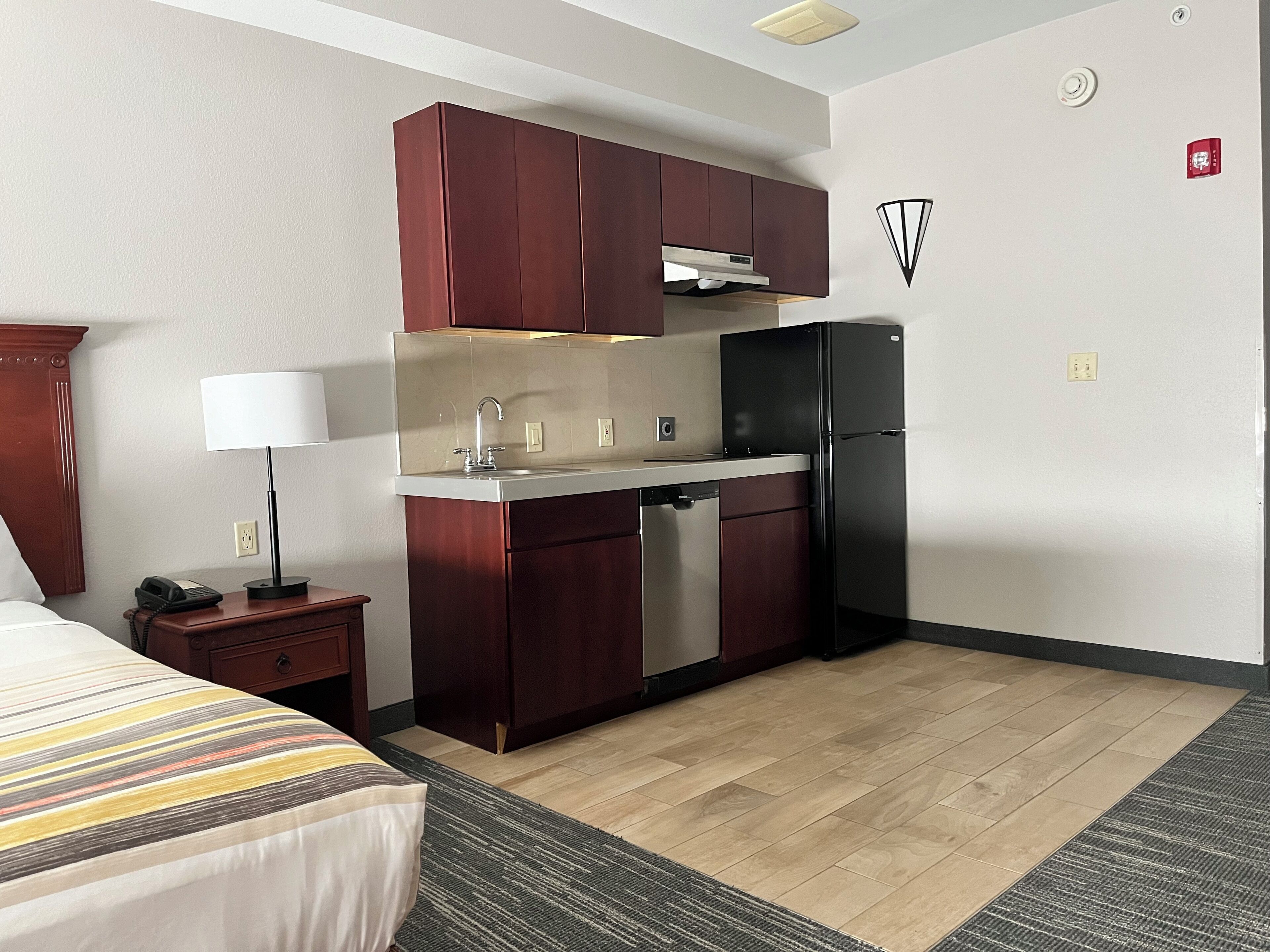 Une chambre d’hôtel de taille réduite comprenant un lit, une petite table de chevet avec une lampe, une kitchenette équipée d’un réfrigérateur, d’un four à micro-ondes, d’un évier et de placards.