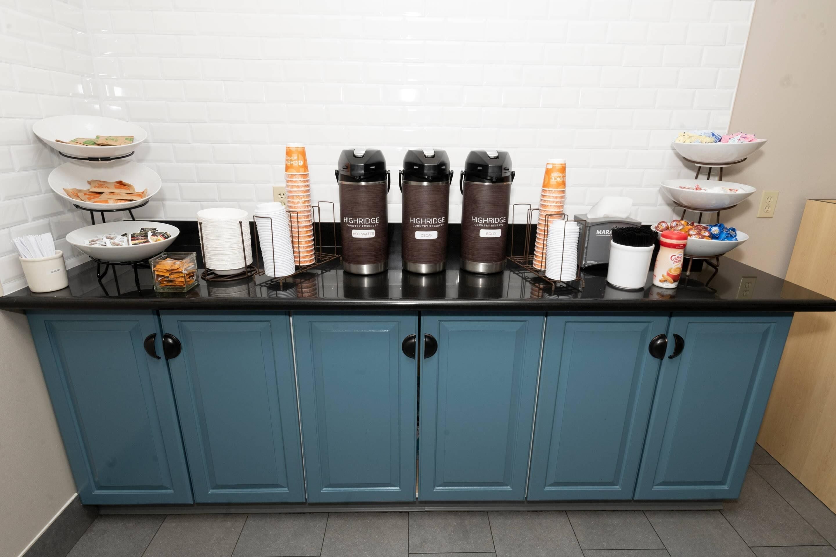 A kitchen area with a blue cabinet, a coffee station with branded dispensers, and various food items on shelves.