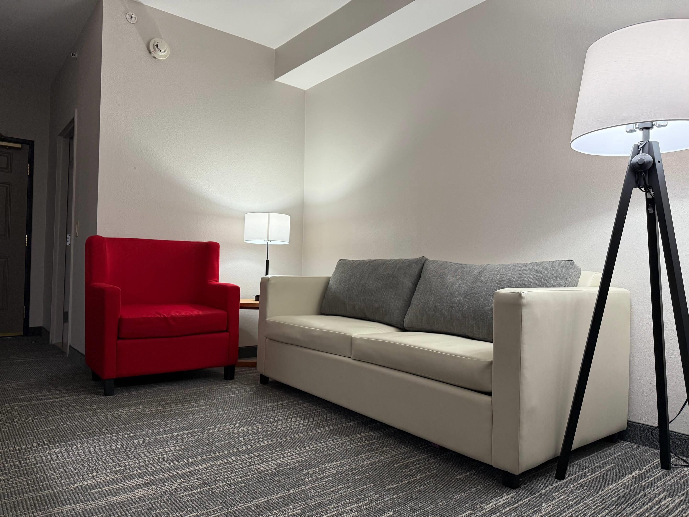 Una habitación de hotel con un sillón rojo, un sofá beige, una lámpara de pie y una mesita de noche.