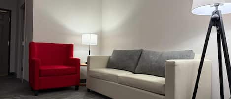 Una habitación de hotel con un sillón rojo, un sofá beige, una lámpara de pie y una mesita de noche.