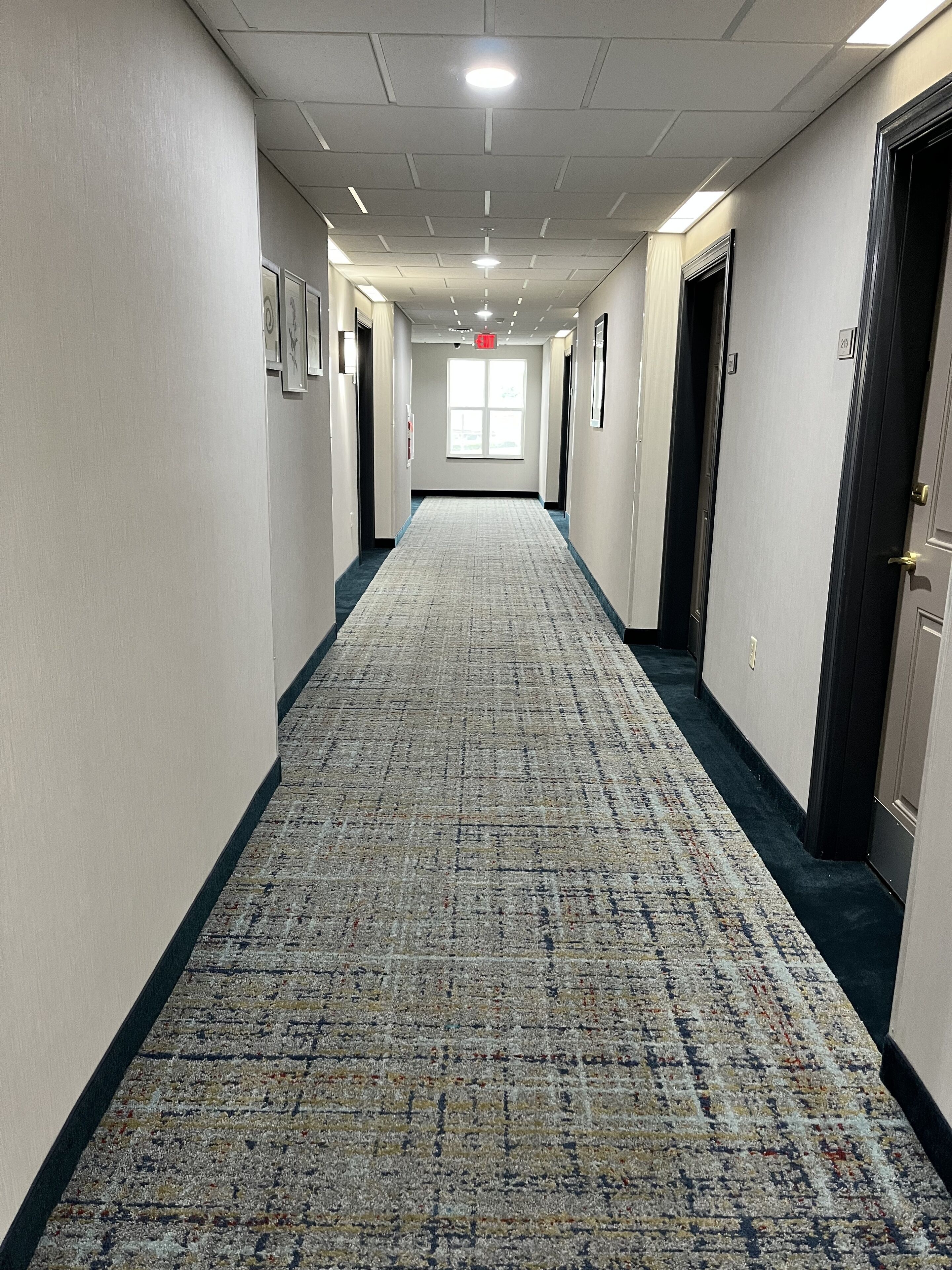 A hotel hallway with a patterned carpet, doors on both sides, and framed pictures on the walls.