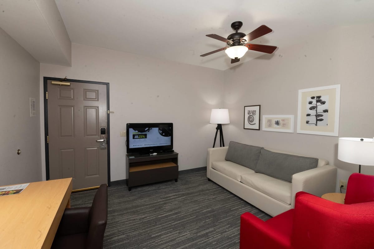 A hotel room with a television, a sofa, a red chair, a ceiling fan, and framed artwork on the wall.