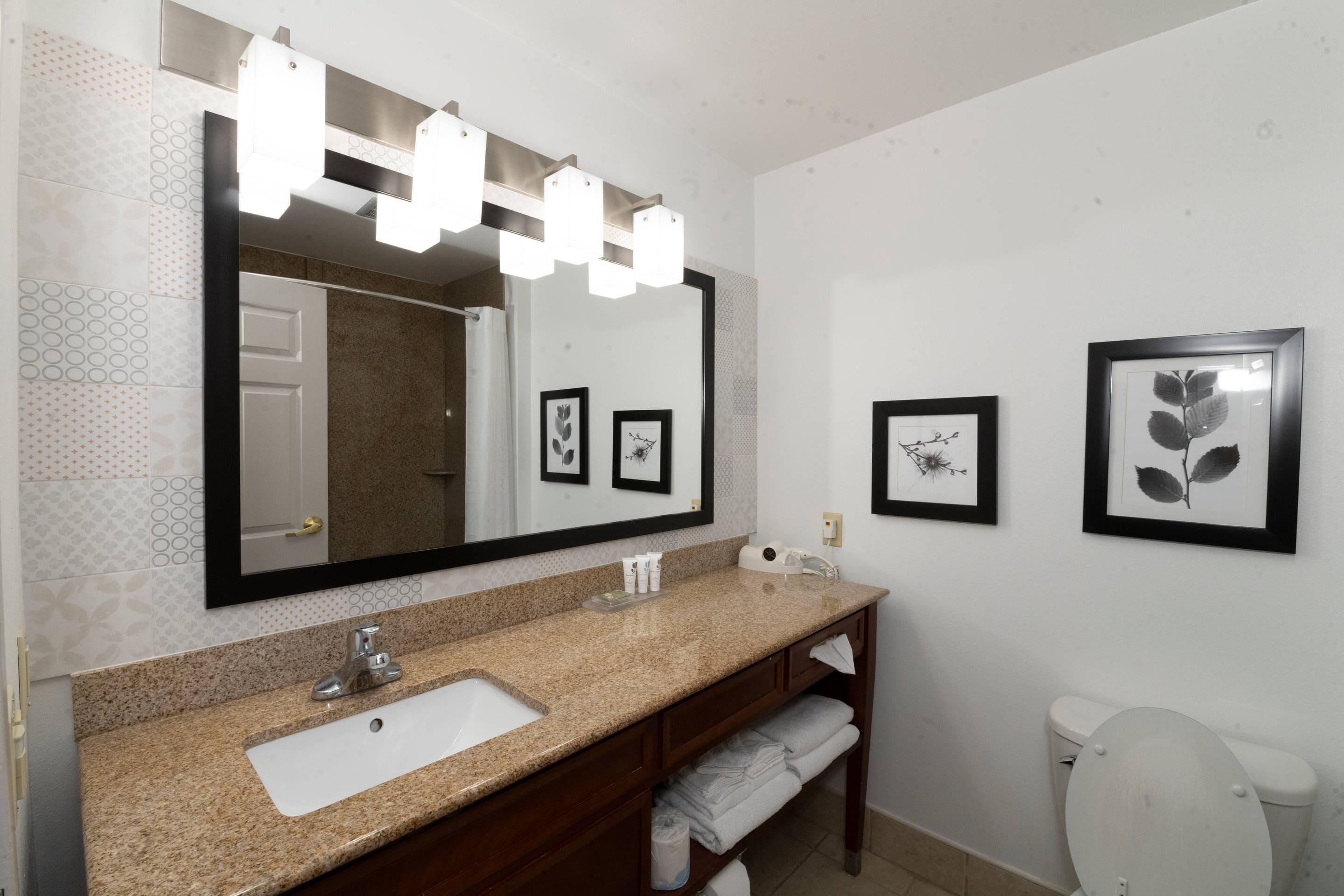 Un baño con lavamanos de granito, un espejo grande con luces, fotografías enmarcadas en la pared y un inodoro.