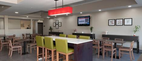 Un comedor moderno con mesas y sillas de madera, una isla central con sillas acolchadas de color verde, una lámpara colgante roja y un televisor de pantalla plana en la pared.