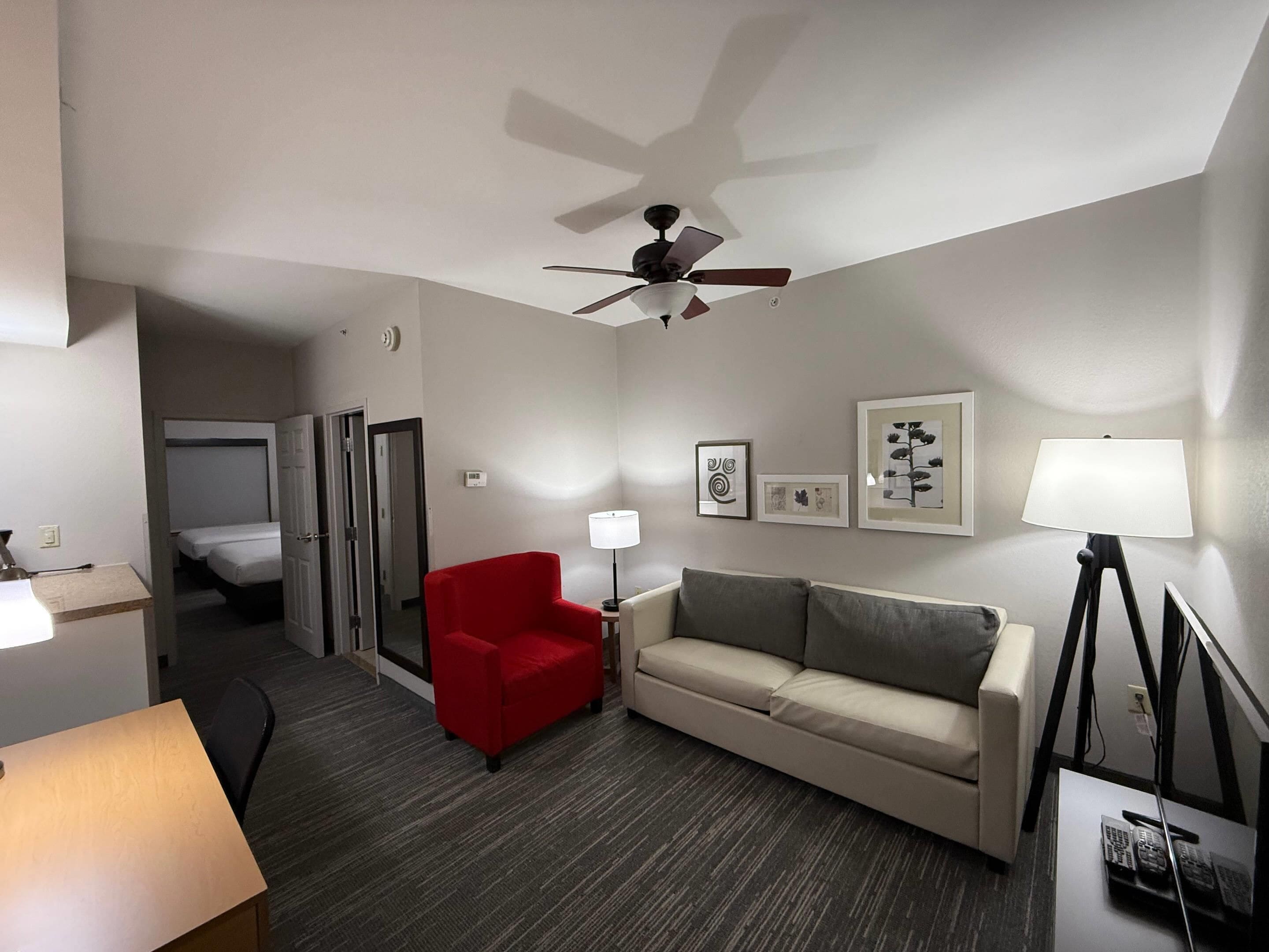 A hotel room with a ceiling fan, a red armchair, a sofa, a lamp, and framed pictures on the wall.