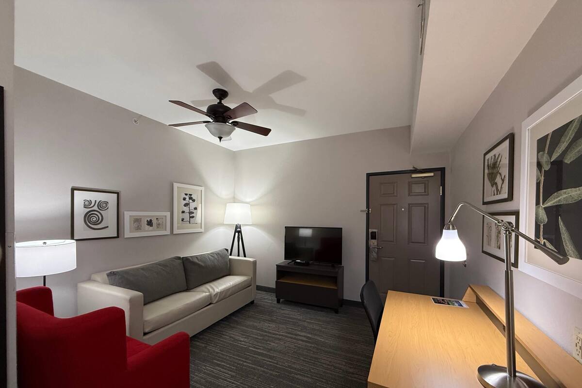 A hotel room with a ceiling fan, a sofa, a red chair, a desk, a TV, and framed artwork on the walls.