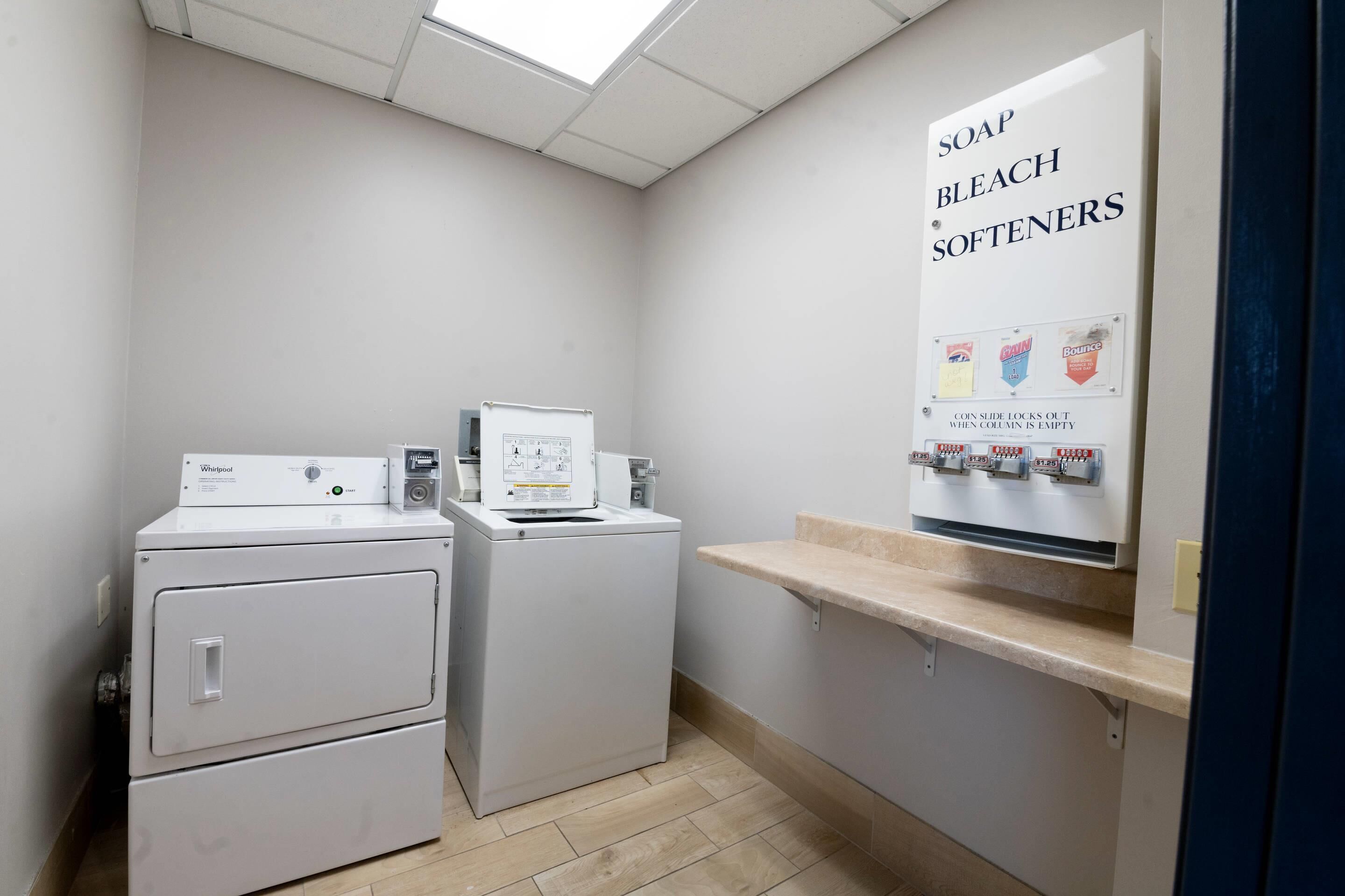 A laundry room with two Whirlpool washers and dryers, a shelf with soap, bleach, and softeners, and a sign indicating the soap dispensers lock out when empty.