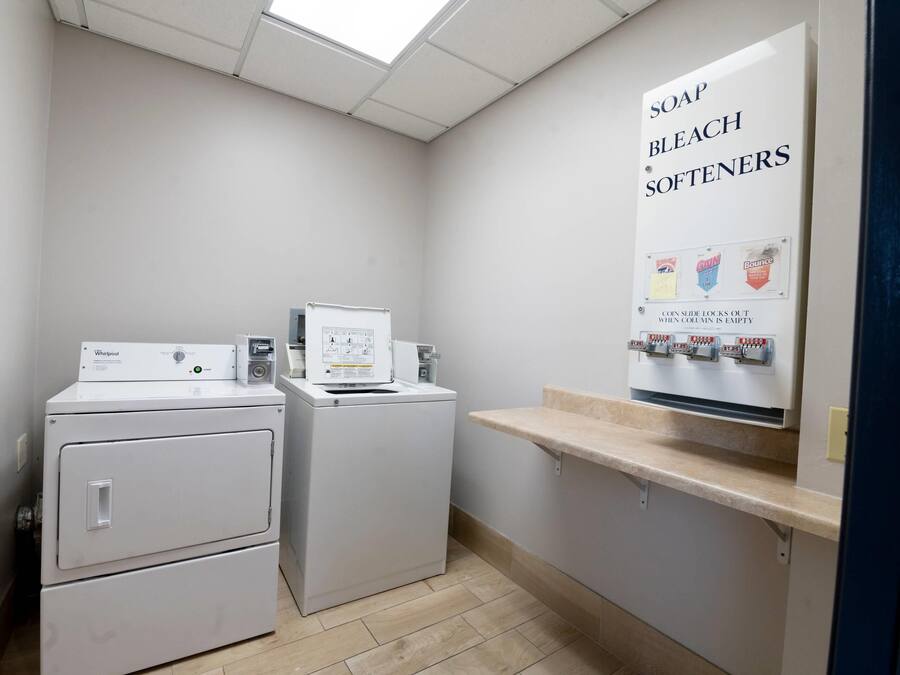 Lavanderia equipada com duas máquinas de lavar e secar Whirlpool, uma prateleira com sabão, água sanitária e amaciantes, e uma placa indicando que os dispensadores de sabão ficam bloqueados quando vazios.