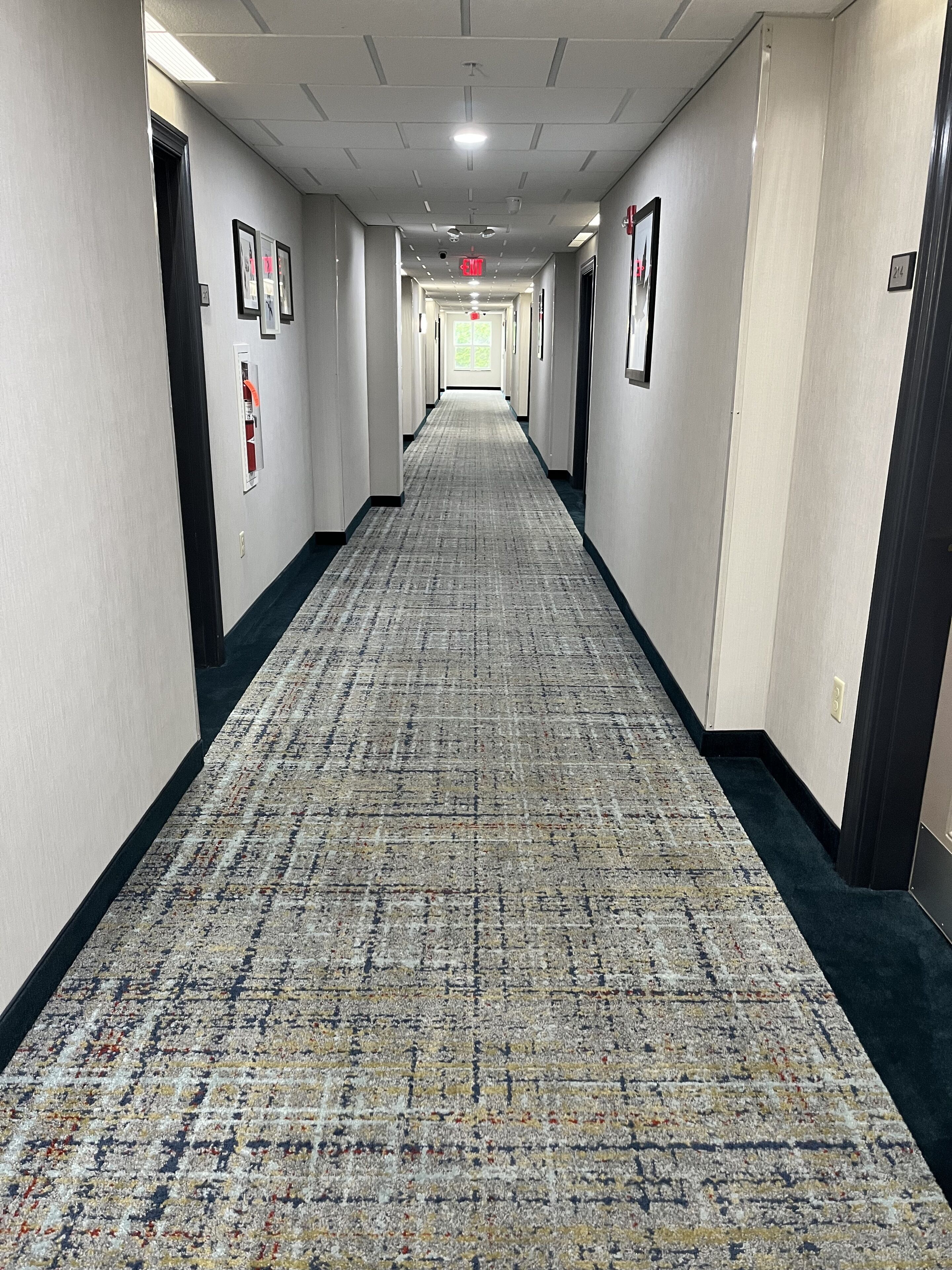 A long, narrow hotel corridor with patterned carpet, framed pictures on the walls, and doors on both sides.