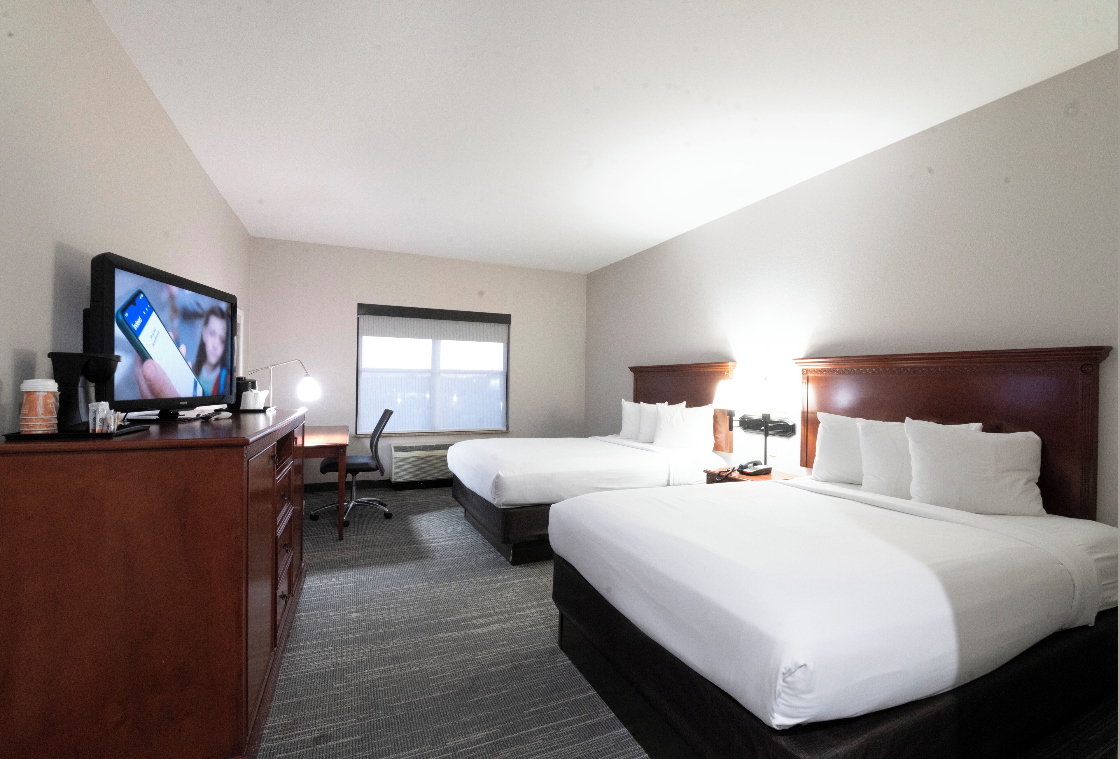 Habitación de hotel con dos camas, un escritorio con televisor y ventana que deja entrar luz natural.