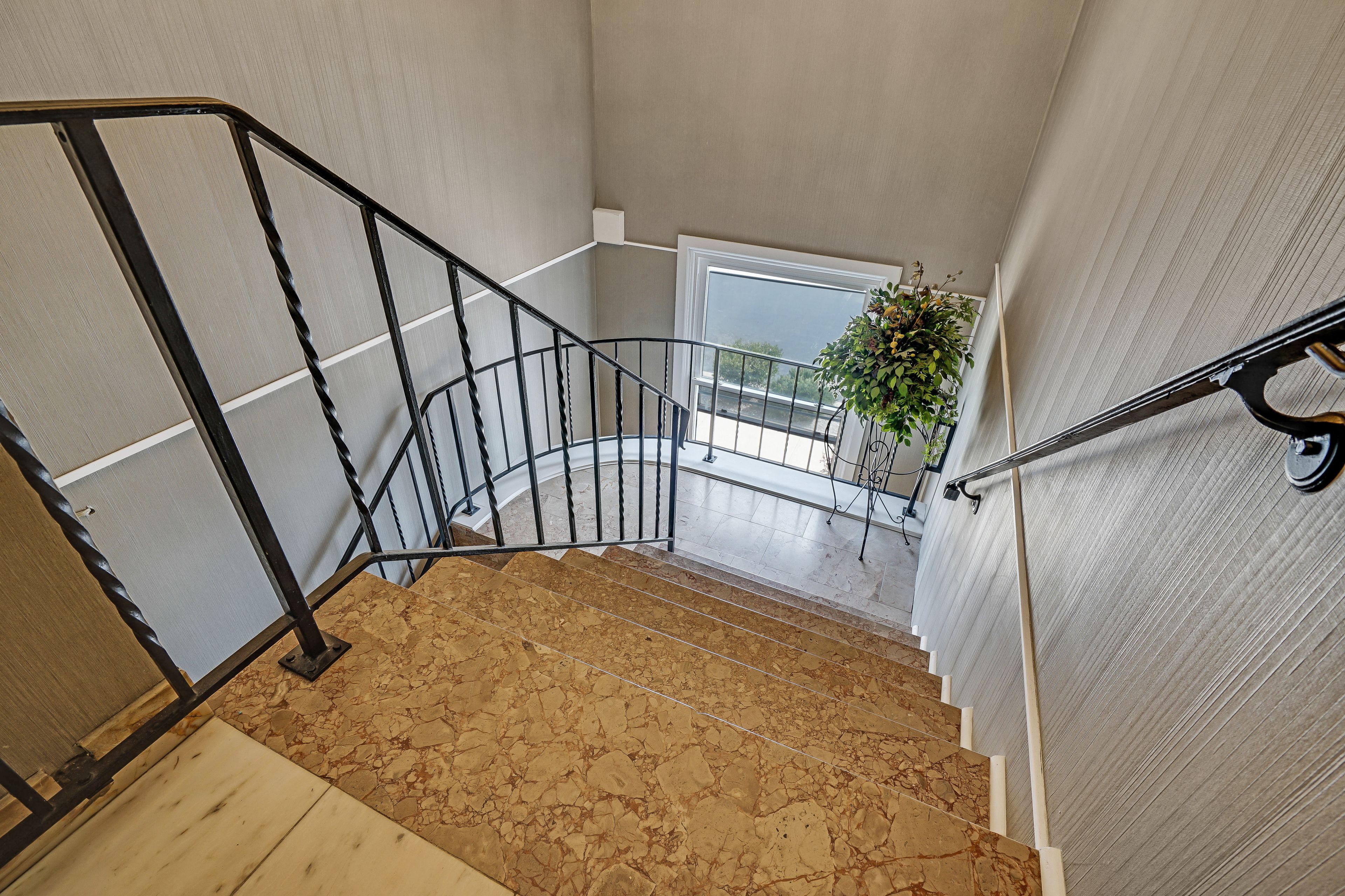 Una escalera con barandilla de metal negro, una planta en maceta y vistas al exterior a través de un ventanal.