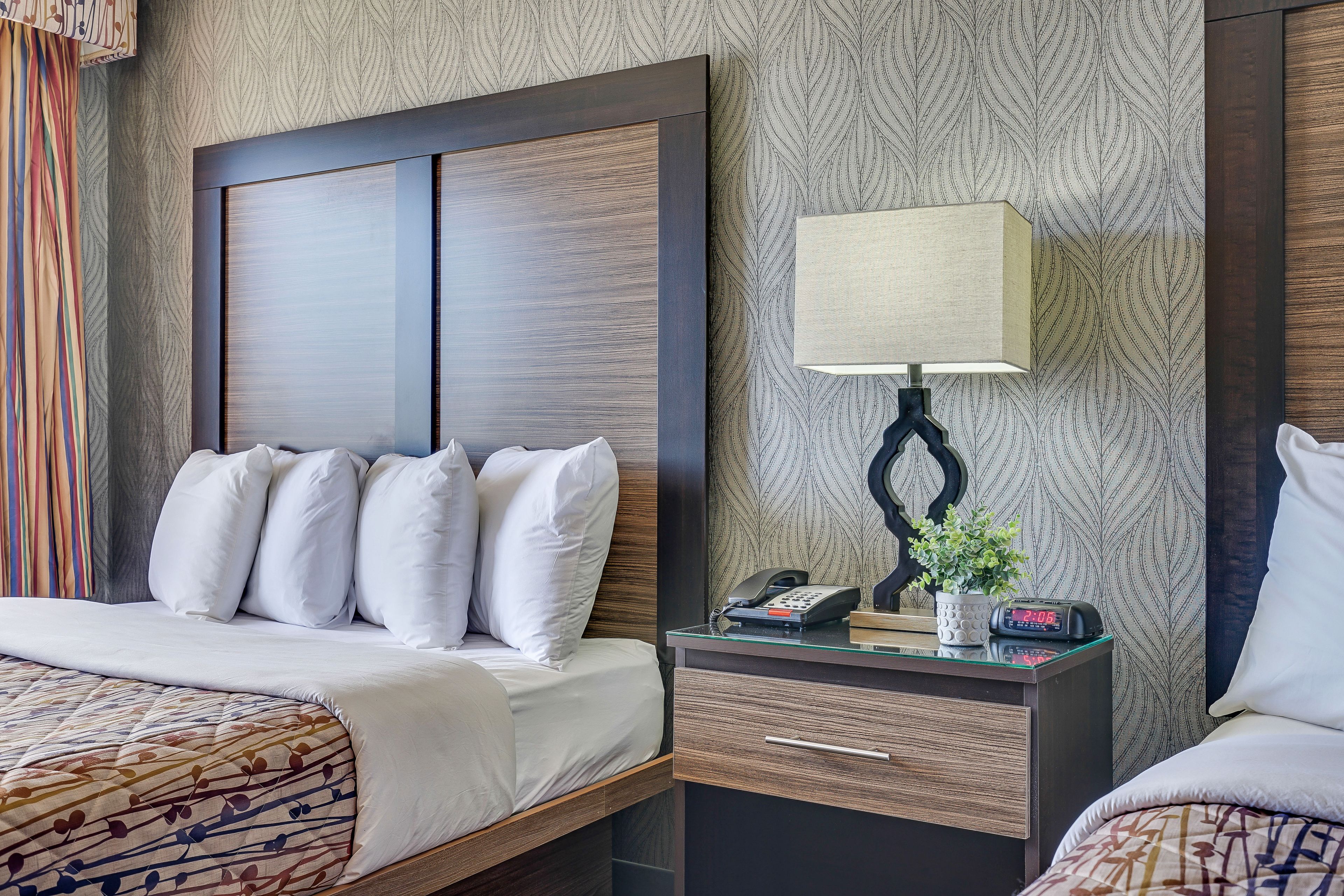 Ein Hotelzimmer mit einem großen Bett, einem Nachttisch mit Lampe, Telefon und einer Uhr.