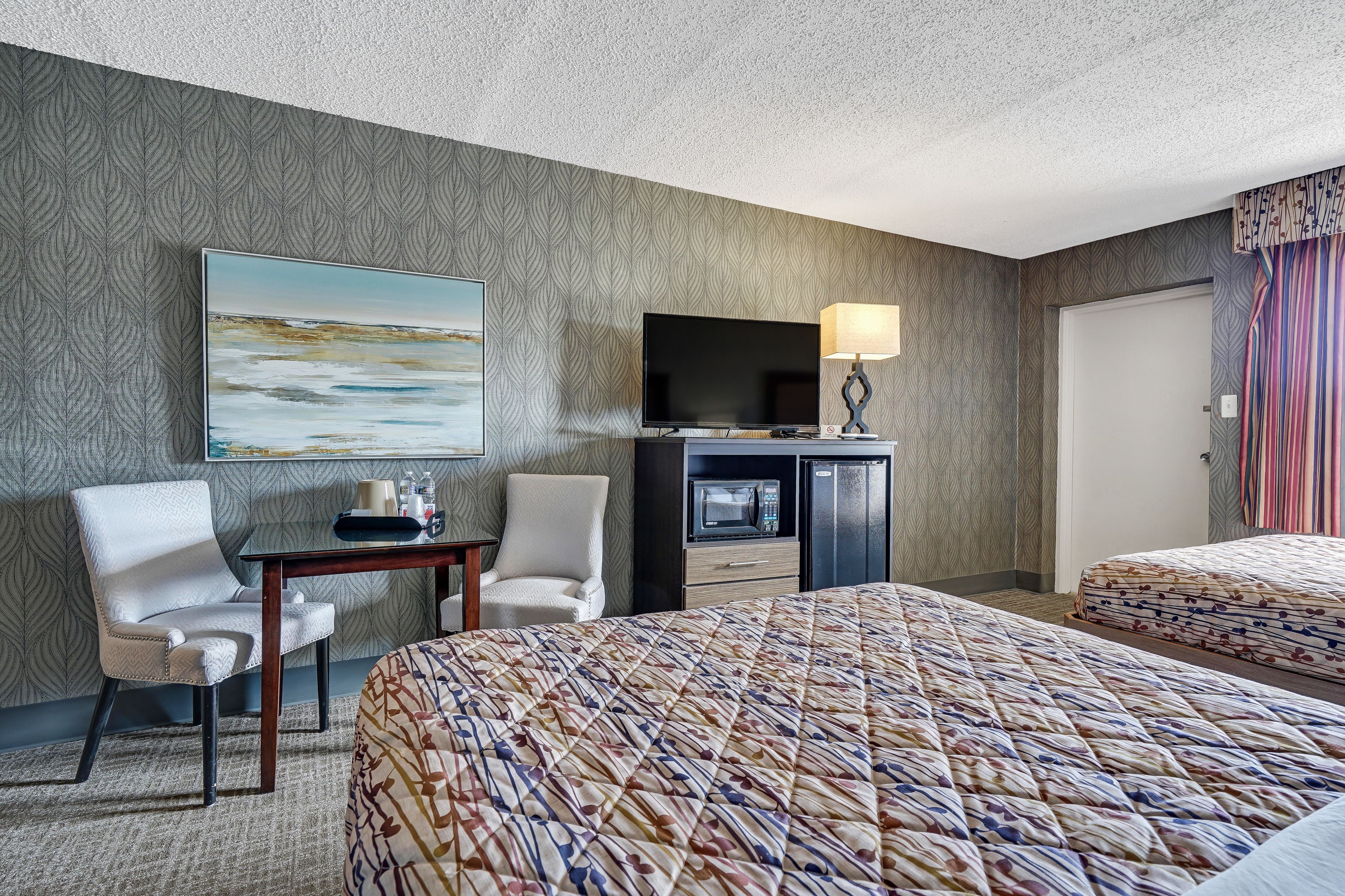 Quarto de hotel com cama, duas cadeiras, escrivaninha com abajur, televisão, micro-ondas e porta.