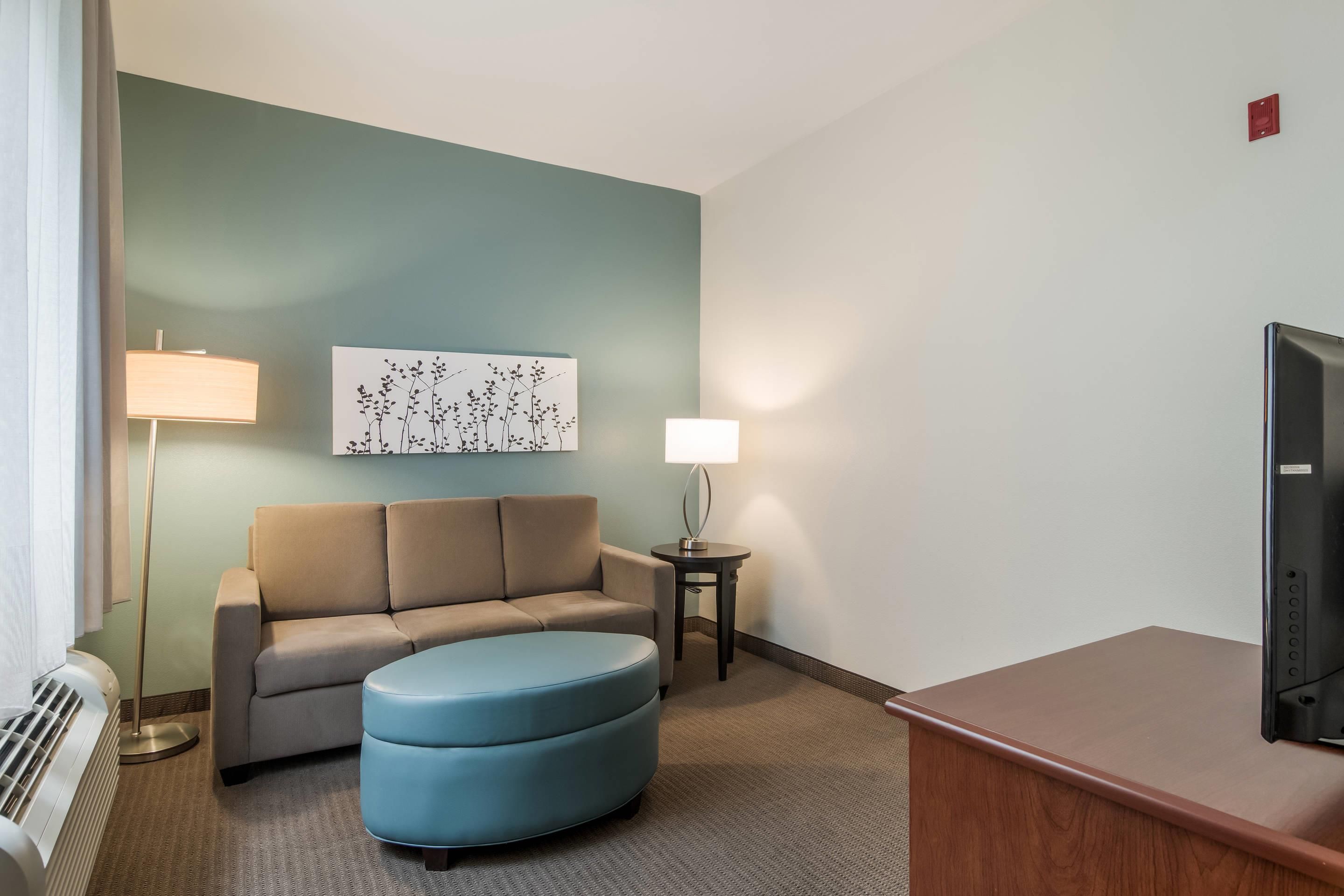 A hotel room with a sofa, ottoman, lamp, artwork, and a television.