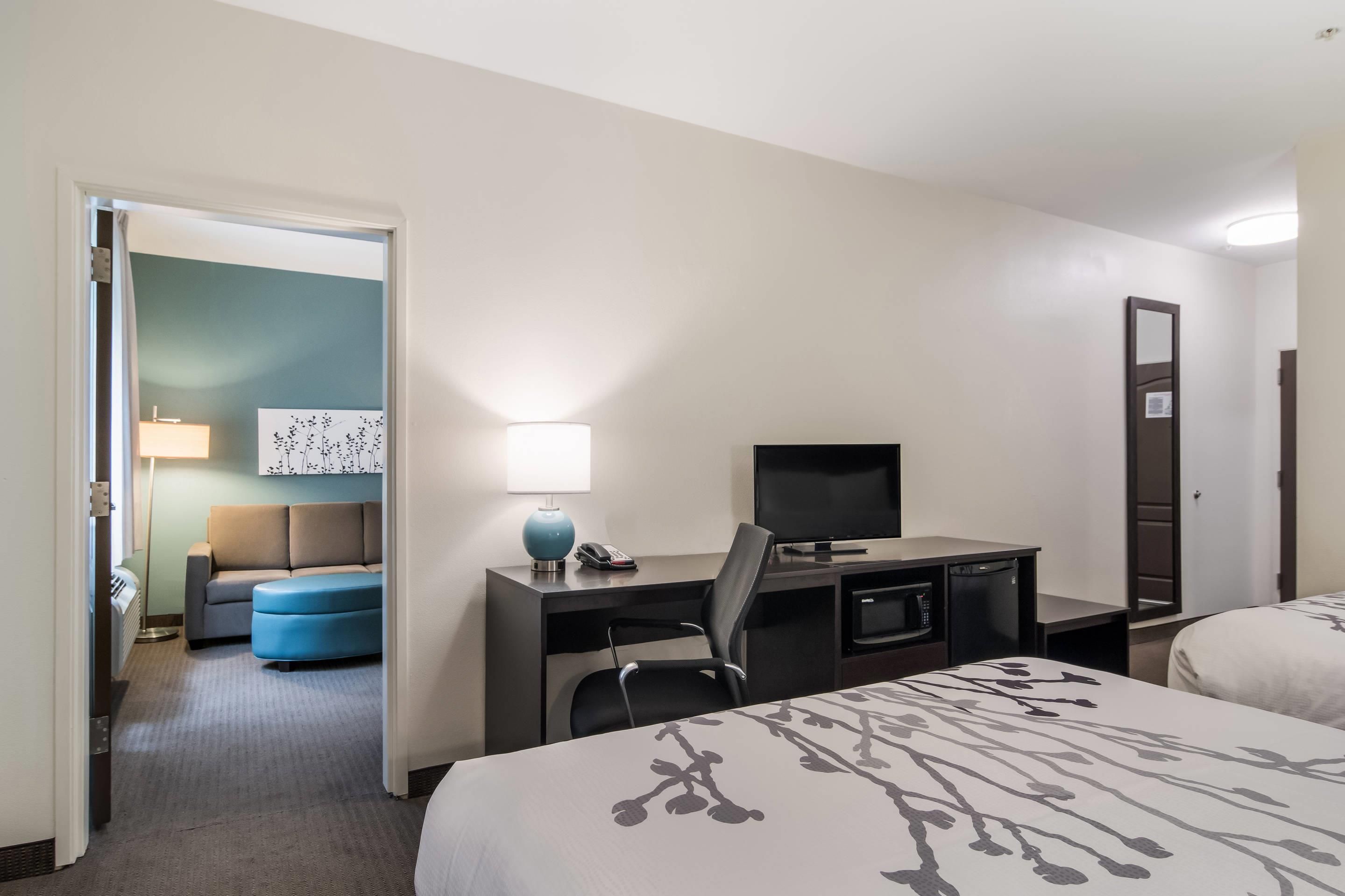 A hotel room with a bed, a desk with a computer, a chair, a TV, and a small kitchenette.