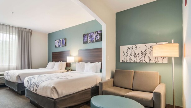 A hotel room with two beds, a sofa, a round ottoman, and artwork on the walls. - Sleep Inn and Suites at Kennesaw State University (Kennesaw)