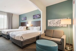 A hotel room with two beds, a sofa, a round ottoman, and artwork on the walls. - Sleep Inn and Suites at Kennesaw State University (Kennesaw)