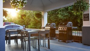 Un patio al aire libre con mesa y sillas, dos parrillas de barbacoa y un arreglo floral colgante.