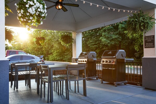 Un patio al aire libre con mesa y sillas, dos parrillas de barbacoa y un arreglo floral colgante.