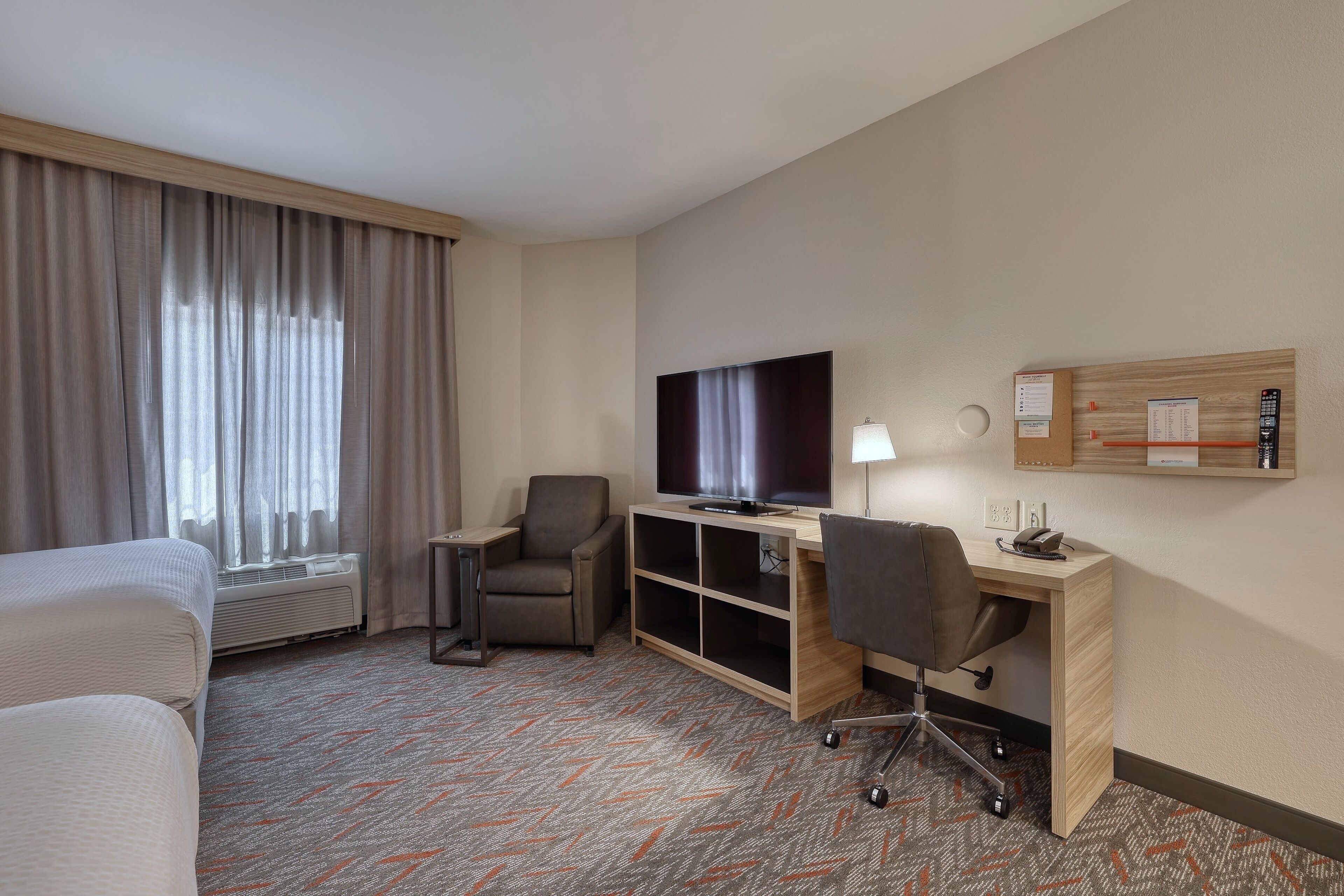 A hotel room with a bed, a desk with a computer, a TV, a chair, and a wall-mounted bulletin board.