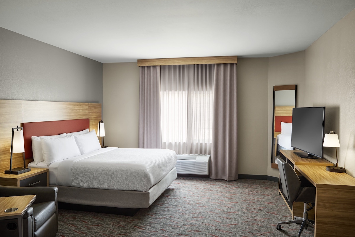A hotel room with a bed, bedside tables, a desk with a chair, a television, and a large window with curtains.
