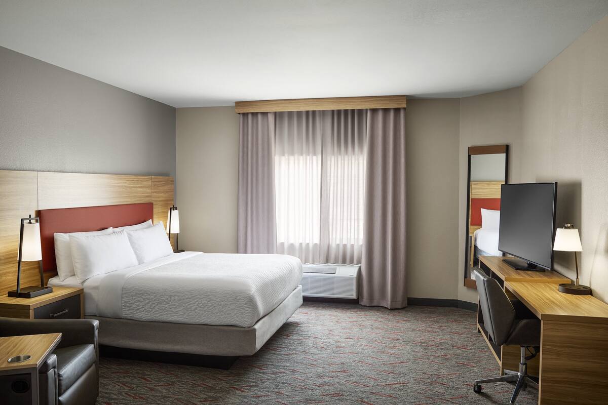 A hotel room with a bed, bedside tables, a desk with a chair, a television, and a large window with curtains.