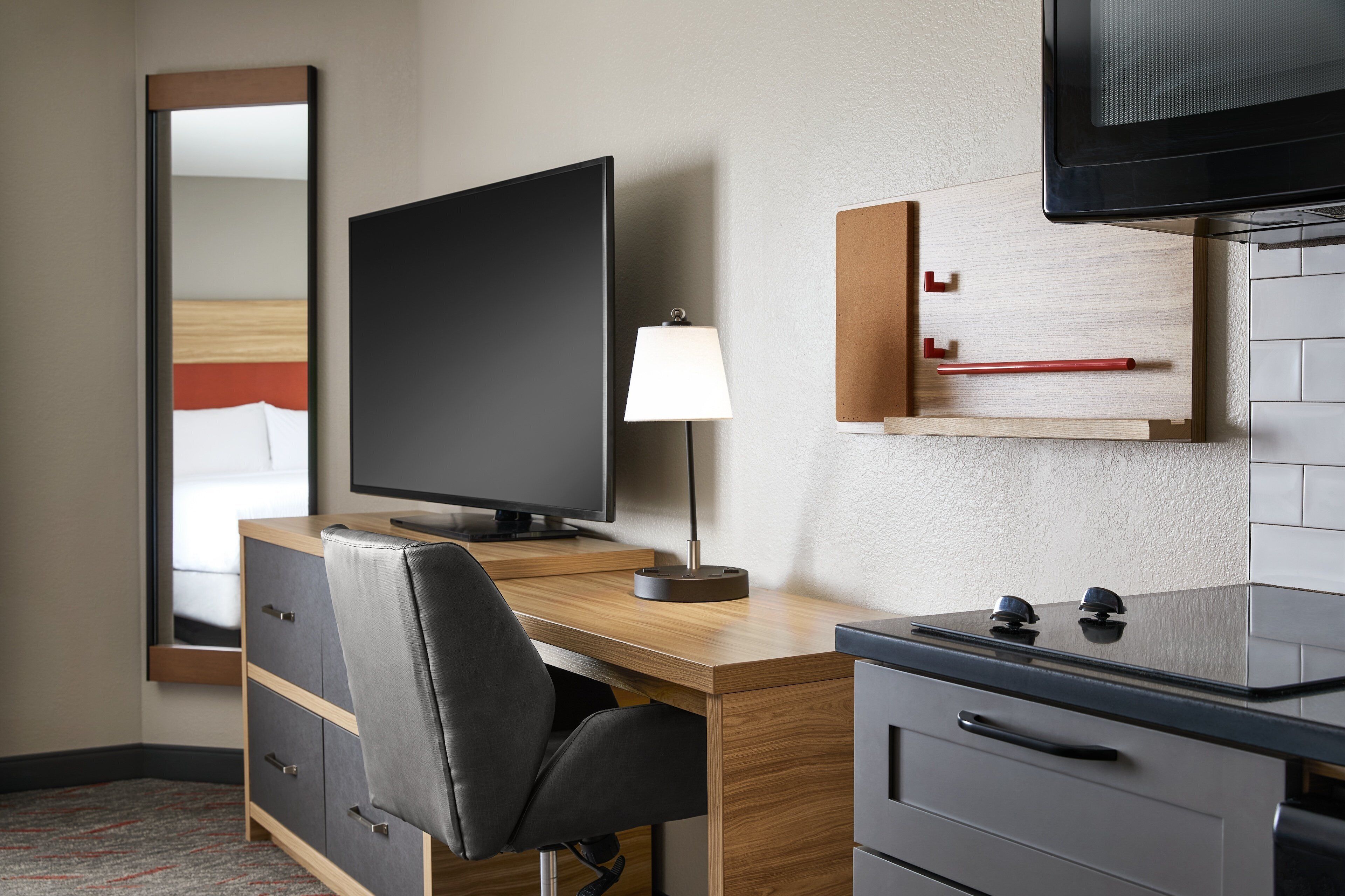 Habitación de hotel con un escritorio de madera, una silla negra, un televisor de pantalla plana, una lámpara, un espejo, un armario y una estufa empotrada.