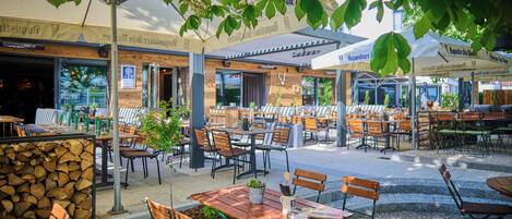 An outdoor cafe with tables and chairs, covered by umbrellas, and a stone wall in the background.