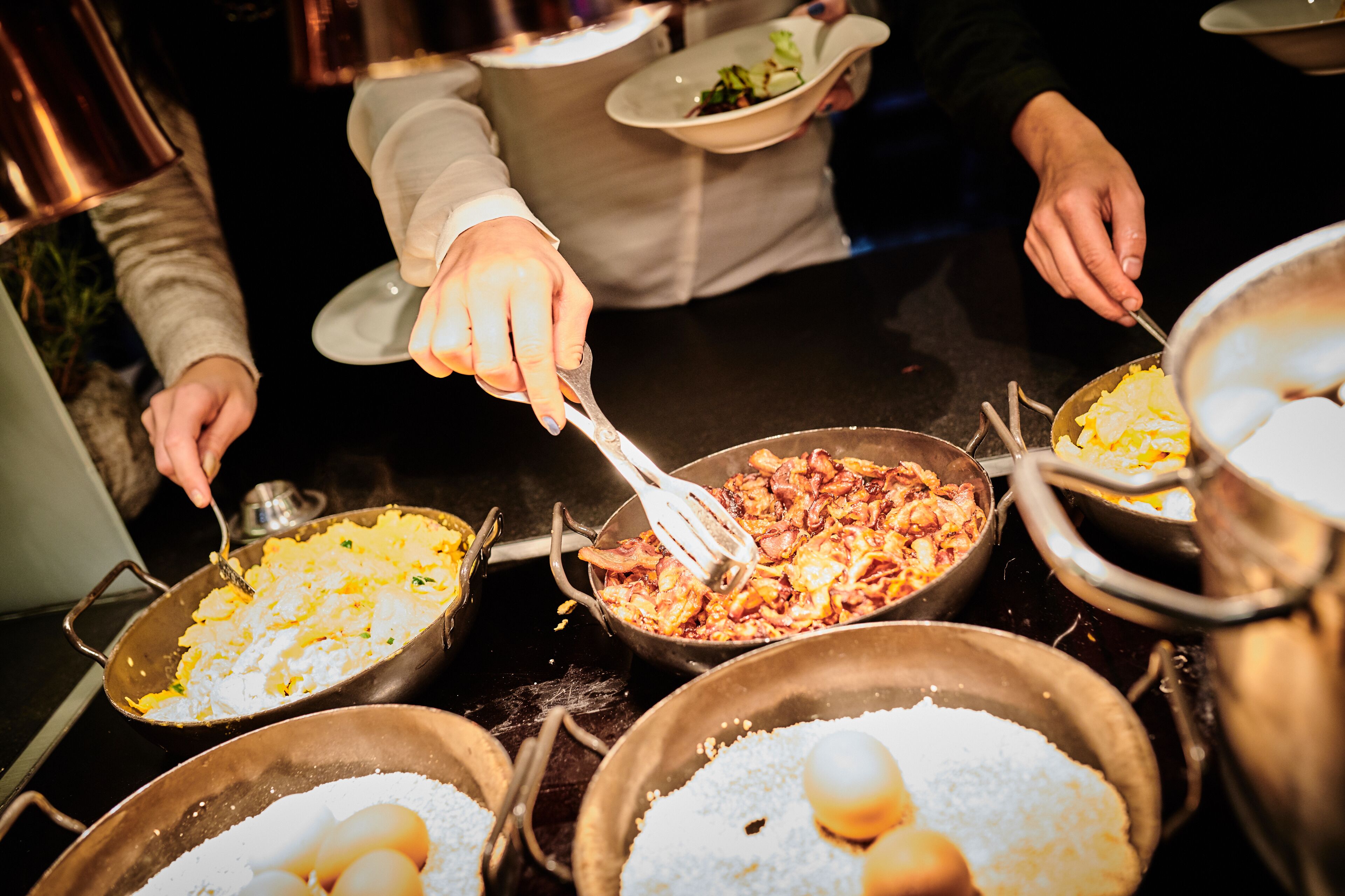 A buffet with various dishes, including scrambled eggs and a meat dish, being served by waitstaff.