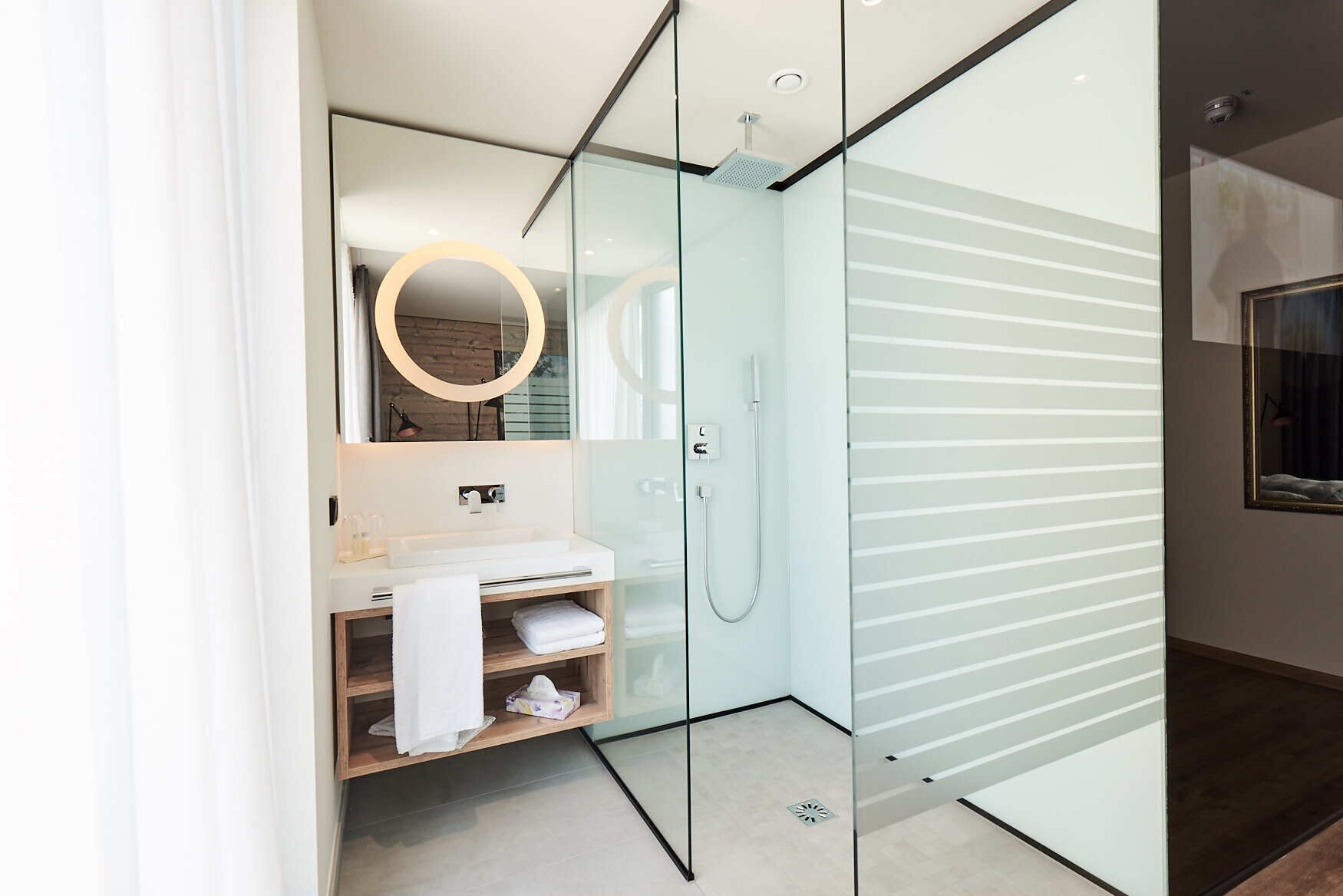 Une salle de bain moderne avec une douche à parois de verre, un meuble-lavabo en bois et un miroir doté d’un éclairage circulaire.