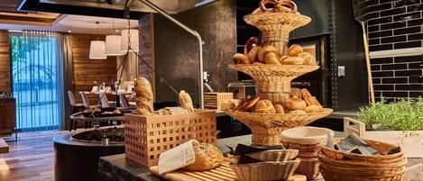 A breakfast buffet with various breads and pastries displayed in baskets, a coffee station, and a seating area with tables and chairs.