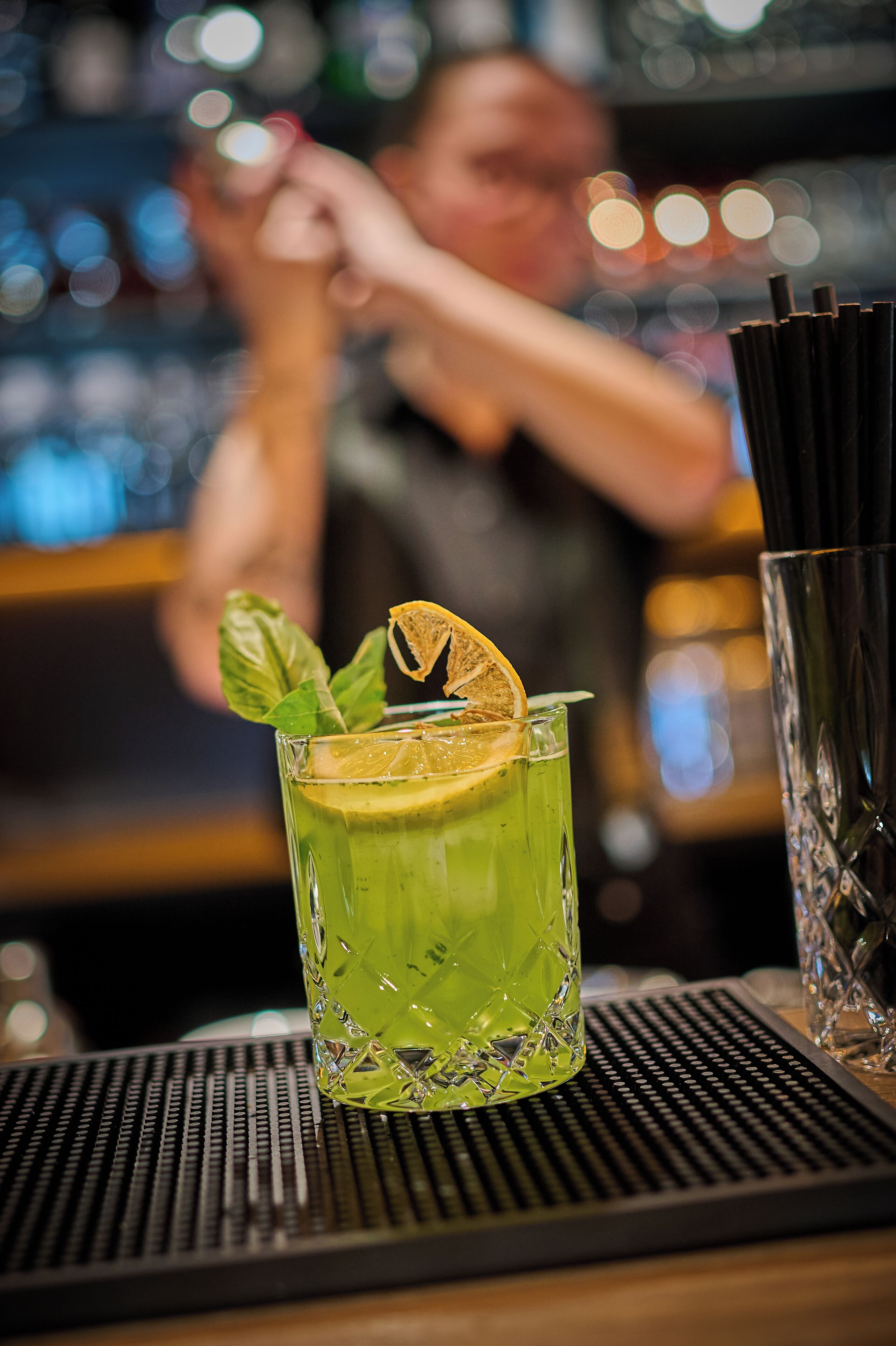 A bar setting with a green cocktail garnished with a lemon slice and mint leaves, a bartender preparing drinks, and a stack of black straws.