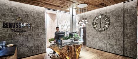 A modern hotel reception area with a wooden ceiling, a large wall clock, and a reception desk with a laptop and decorative plants.