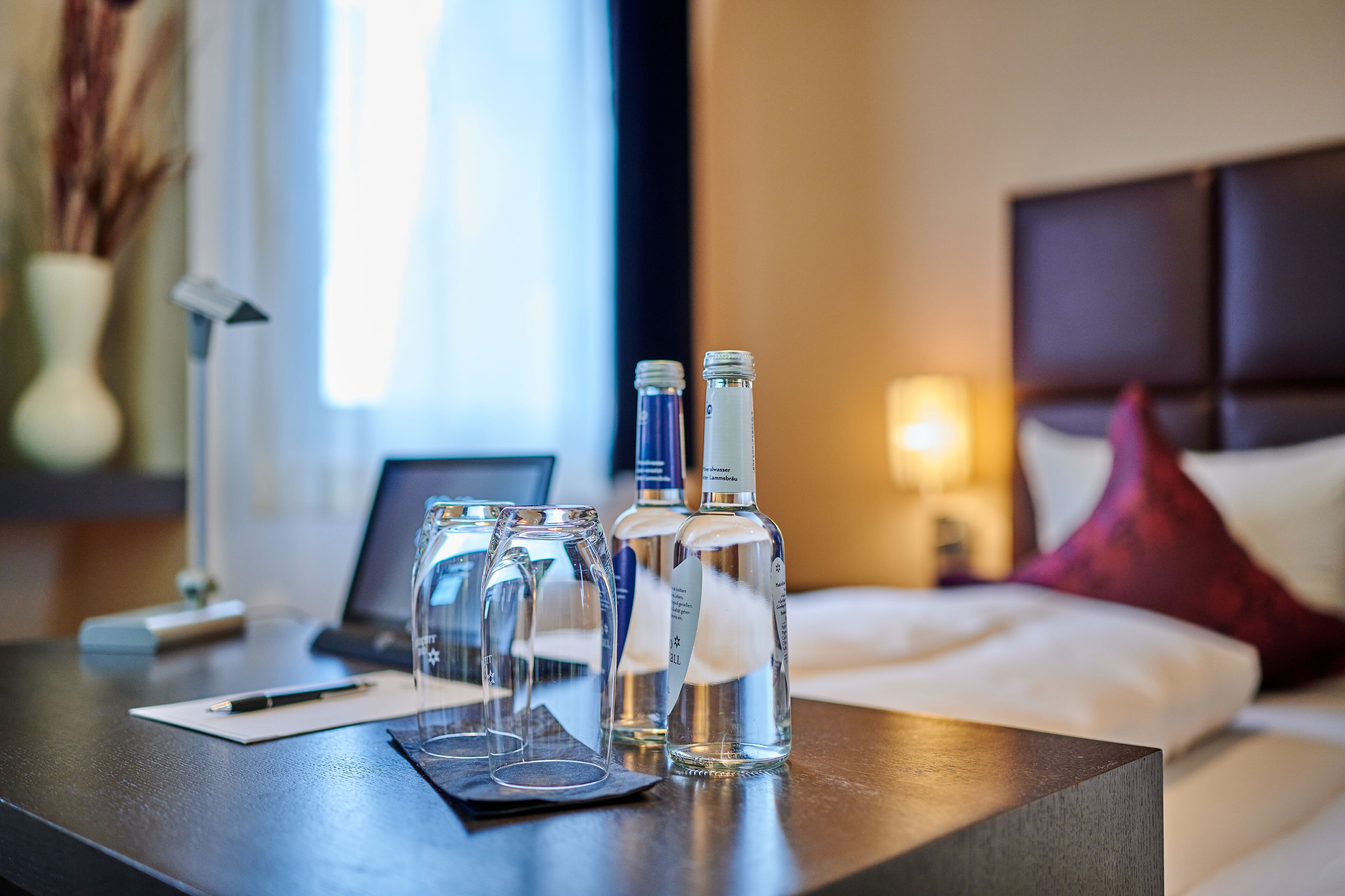 Hotellihuoneessa on puinen yöpöytä, jonka päällä on kannettava tietokone, kynä ja kaksi vesipulloa.
