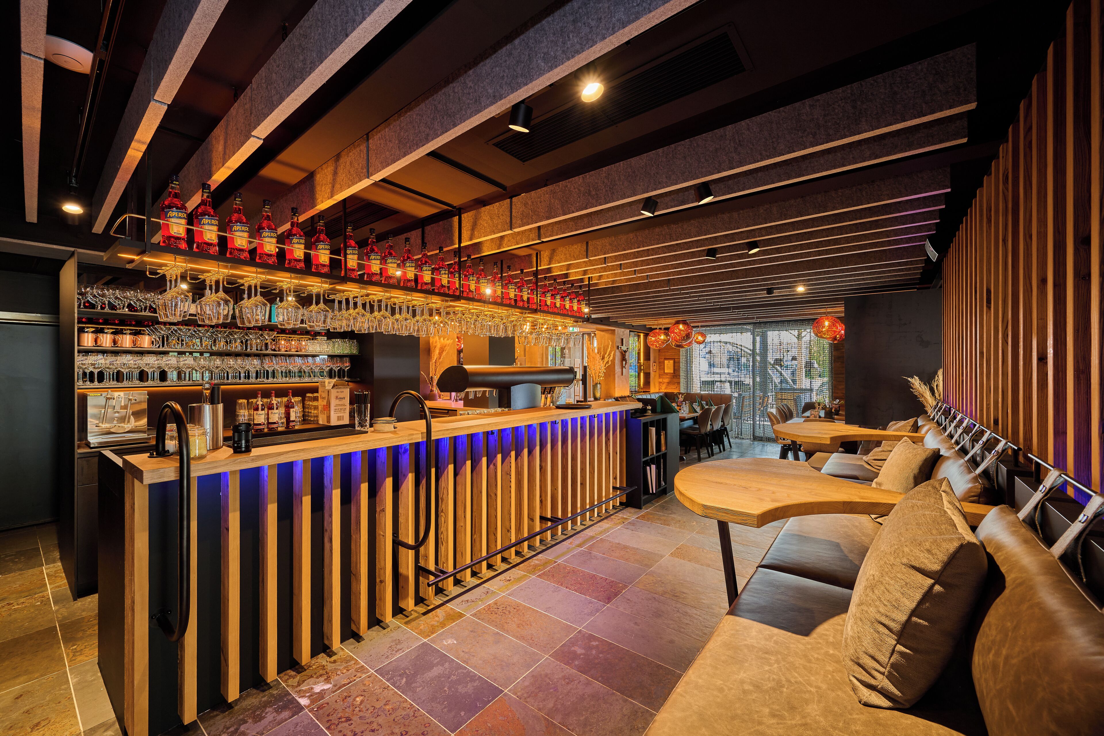 A bar area with a wooden counter, shelves of bottles, glasses, and a seating area with cushioned benches.