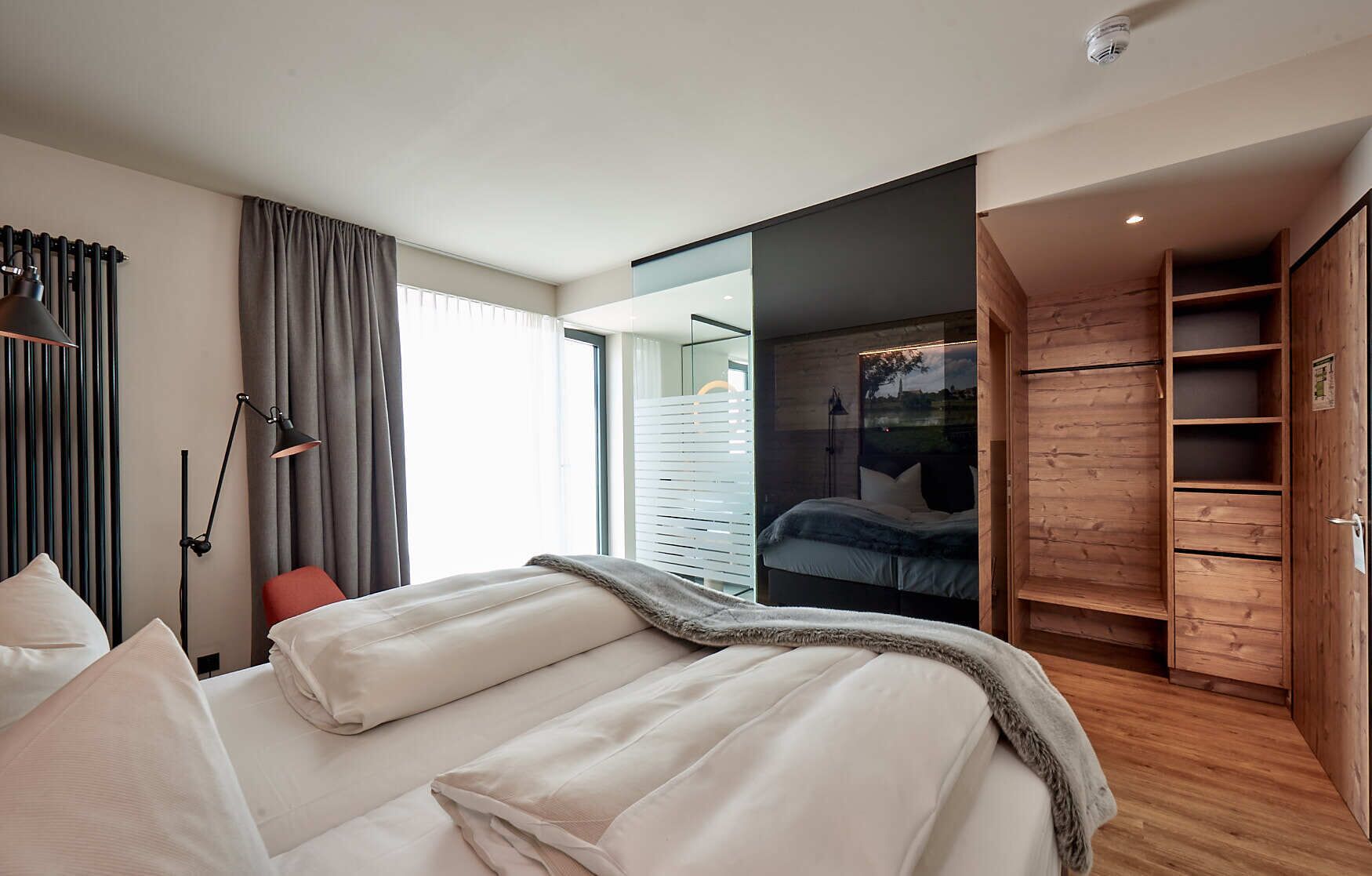 Quarto de hotel moderno com uma cama grande, um guarda-roupa de madeira e vista através de um painel de vidro.