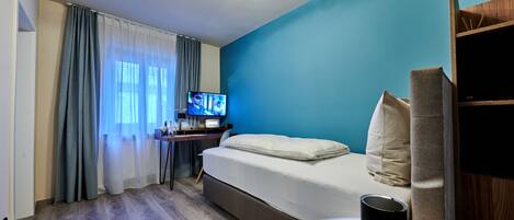 A modern hotel room with a bed, a desk with a computer, a TV, and a window with curtains.