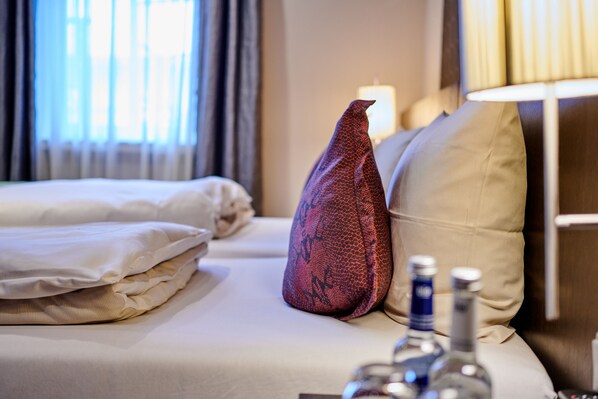 Una cama bien hecha con sábanas blancas, un cojín decorativo rojo y una botella de licor al lado.