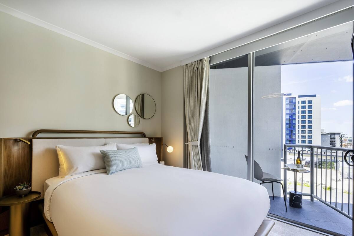 Camera d'hotel con un letto grande, un comodino con una lampada, un tavolino con una pianta e vista sulla città da un balcone.