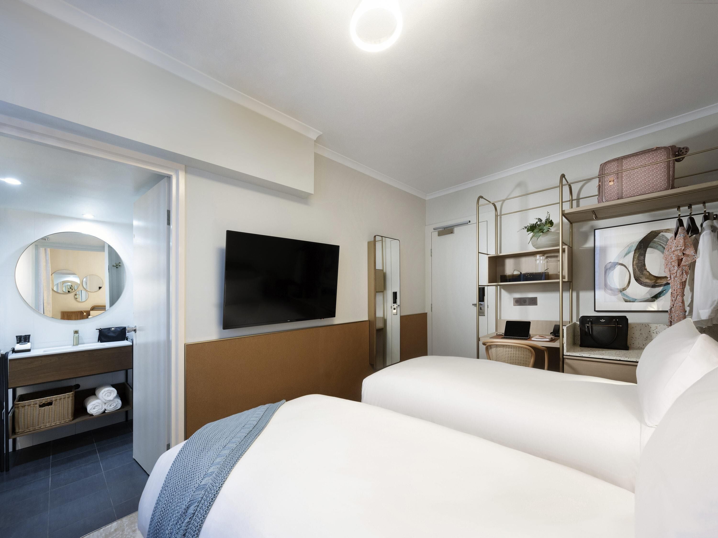 A modern hotel room with a flat-screen TV, a large bed, a desk with a laptop, a mirror, and a wardrobe with hanging clothes.