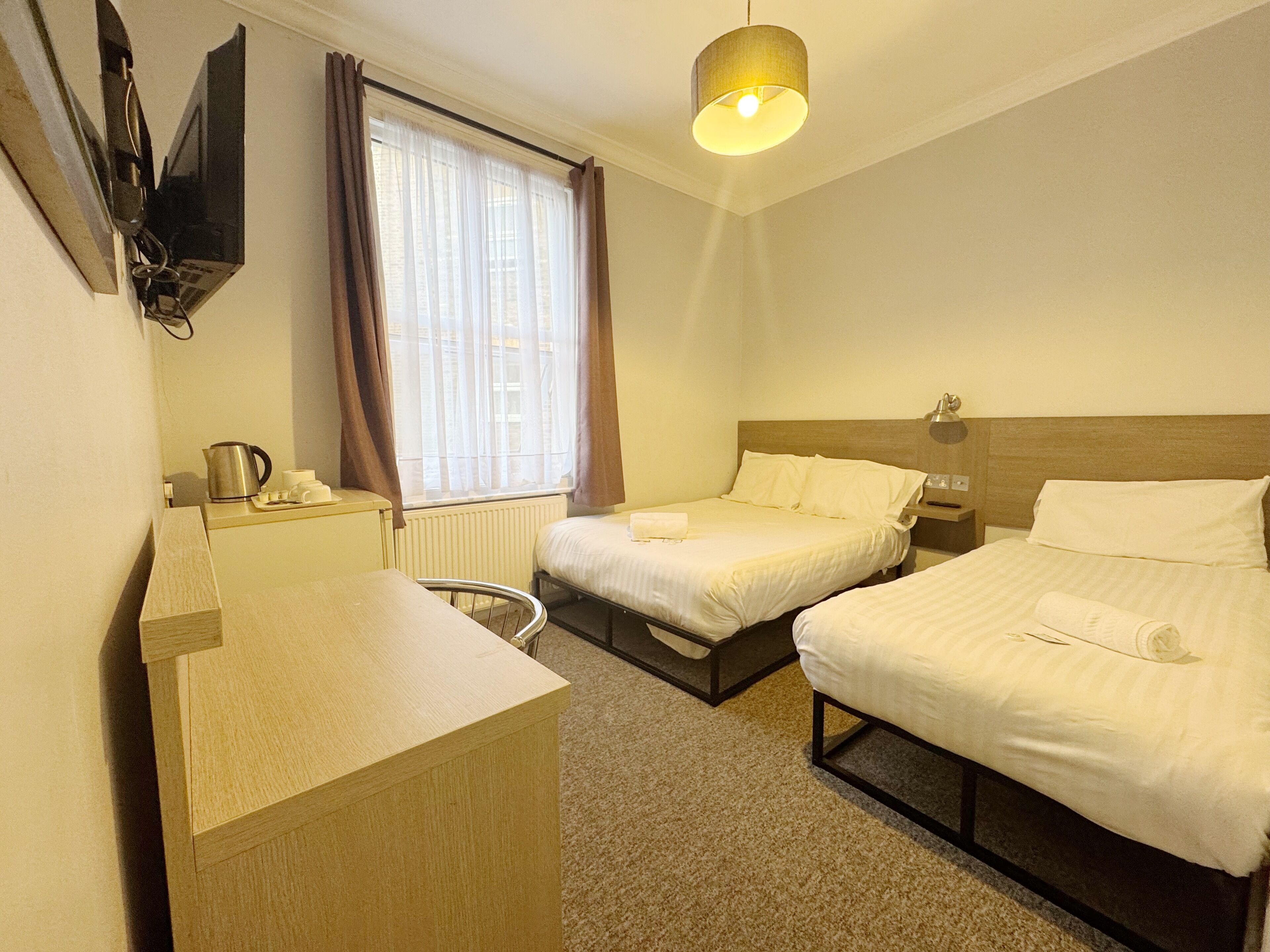 A hotel room with a bed, a desk, a chair, a window with curtains, a radiator, and a wall-mounted TV.