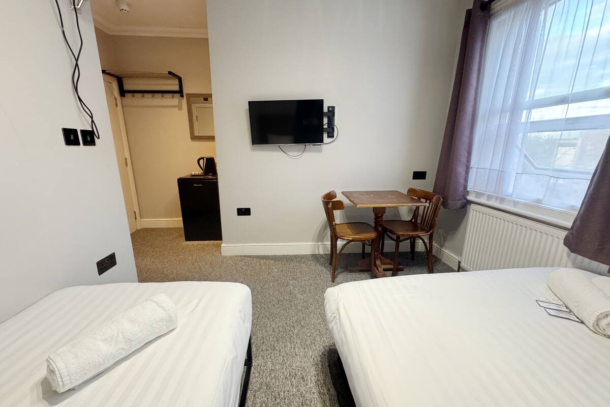 A hotel room with two beds, a TV, a small table with chairs, a heater, and a window with curtains.